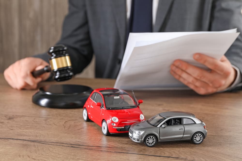 Auto accident law concept: A lawyer sits at a wooden table indoors with documents and a gavel, while the focus is on toy cars in the foreground.