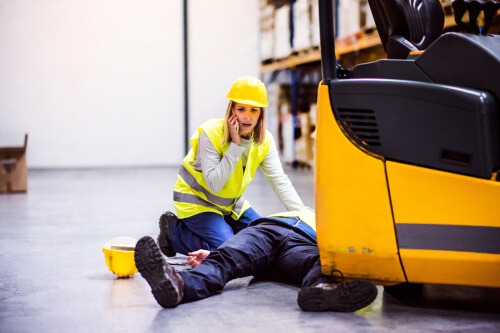 worker laying on ground with a forklift on top of his leg, a fellow coworker is calling for help in the background