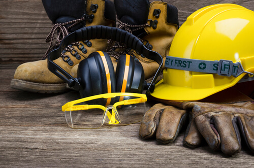 work boots, earmuffs, safety goggles, hard hat and gloves assembled together on a wood surface