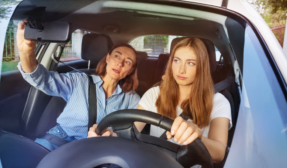 mom teaching teenage daughter proper driving techniques by adjusting the rearview mirror and providing a space free of distractions