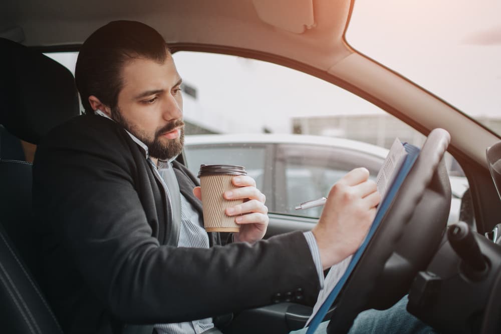 man driving distracted while looking over notes and writing with a cup of coffee in one hand and talking on the cellphone