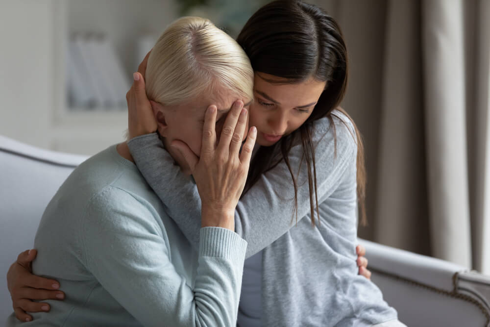 Empathic young lady embracing soothing crying depressed elder mommy, sitting together at home.