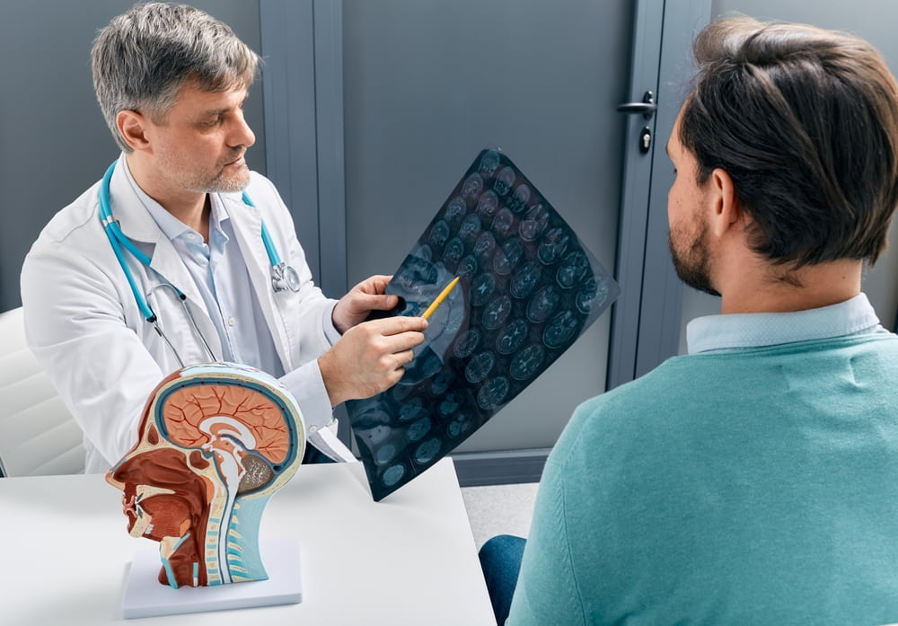 doctor holding brain xray images talking with male patient at a desk with a human skull model sitting on it