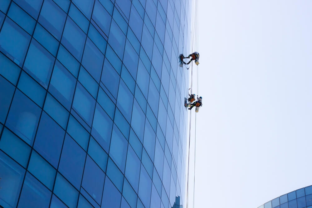 Window cleaning workers hanging outside blue glass office building.