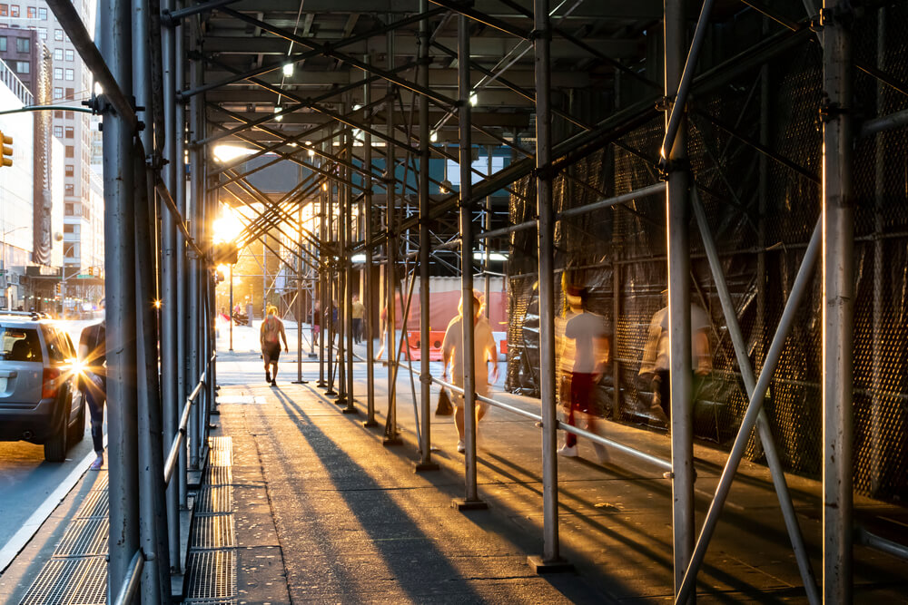 People walking under construction scaffolding on the streets of New York City with the bright light of sunset shining in the background.