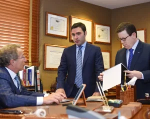 Three men in business suits are discussing documents in a law office, with framed certificates on the wall behind them.