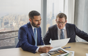 Two men in business suits sit at a conference table having a discussion, with a city skyline visible through the window behind them.