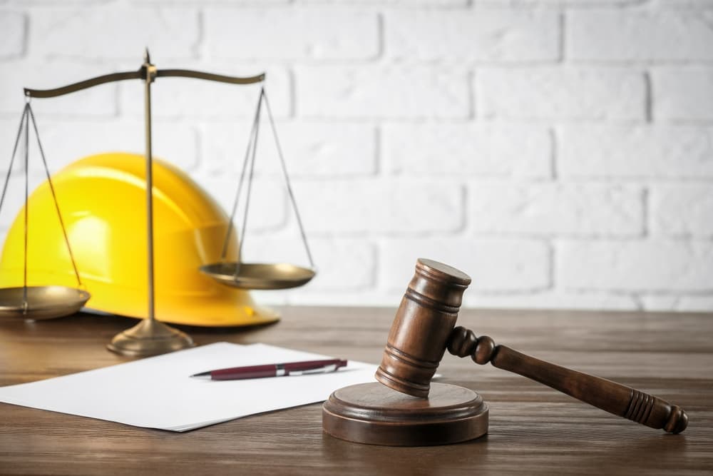 Labor and construction concepts represented: judge's gavel, scales of justice, protective helmet, and a document with a pen on a wooden table.