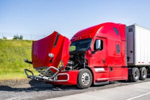A red, broken-down big rig semi-truck with its hood open and an attached dry van trailer sits out of service on the road shoulder, waiting for a tow truck or mobile roadside assistance.